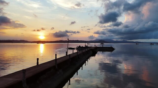 Aerial drone view of a concrete fishing jetty at sunset with a fishing boat moored alongside, over a golden glassy bay with mountain ridges, Taytay, Palawan, Philippines.