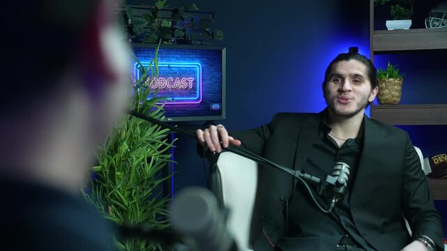 Podcast guest speaking into microphone in blue lit recording studio, host blurred in foreground