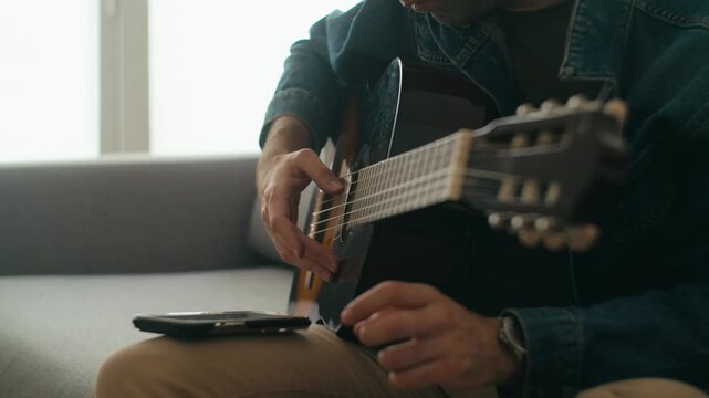 A man in a denim jacket sits on a sofa playing a classical acoustic guitar, using a smartphone for chords or recording. Authentic scene for music education and wellness content.
