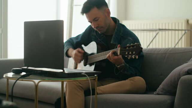 An authentic scene of a young man practicing acoustic guitar at home using a laptop for remote learning or digital music lessons.