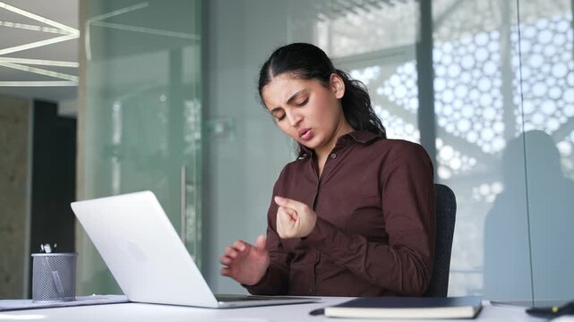 Tired businesswoman suffering from wrist pain while using laptop at workplace in modern business office. Sad female employee massages a joint, does an exercise with her hand.