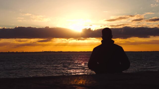 Silhouette person watching golden sunset over a rough sea as clouds drift across the evening sky