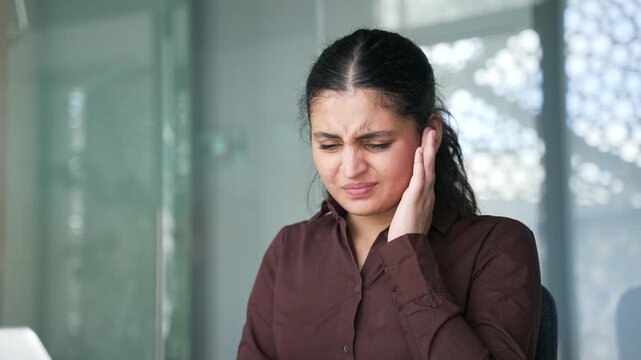 Businesswoman suffers from ear pain sitting at workplace in office. Female touches her ear with her fingers. She has furuncle, shingles, eczema, ear diseases or otitis media. Earache. Close up
