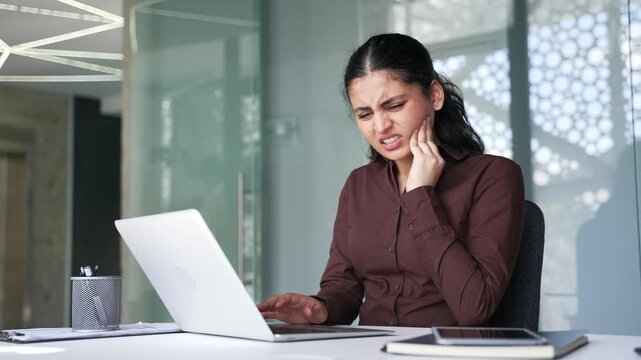 Upset businesswoman suffering from toothache while sitting at workplace in business office. Woman employee holds her hand near her cheek, massaging a sore spot. She needs help from the dentist