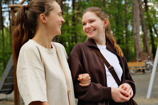 Two friends stand close to each other in a park. They share smiles while enjoying time outdoors. Trees surround them, and the atmosphere is lively and bright