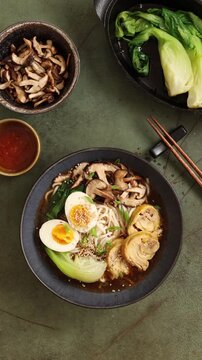 Top view vertical video of hand sprinkling sesame seeds over ramen soup. Final garnish stage of Asian noodle dish preparation on dark flat lay background.