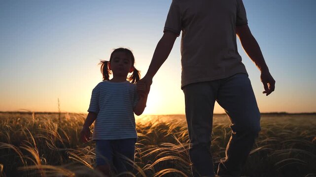 A man walks hand-in-hand with a girl in a field during sunset. The silhouettes of the man and girl create a peaceful scene against the sunset sky.
