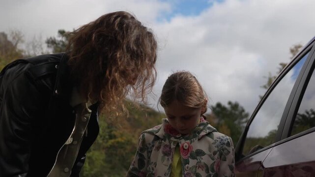 Mother daughter travel: Loving adult woman zipping up little girl jacket near car on family road trip.