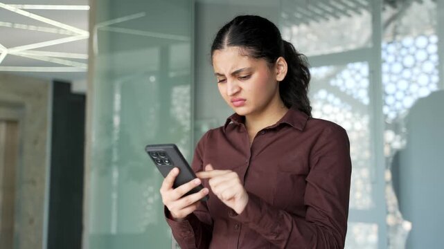 Angry young businesswoman having issues with using mobile phone app at workplace in business office. Confused female feeling annoyed with low battery, problems with smartphone or unstable connection.
