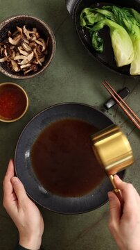 Top view vertical video of pouring hot ramen broth into a bowl. Asian noodle soup preparation on dark background with chopsticks, pak choi, shiitake mushrooms, chili sauce and cooking ingredients.