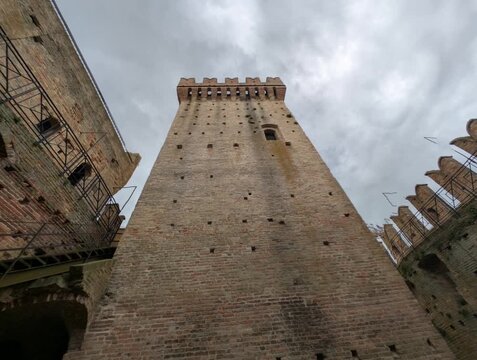 Torre medievale in mattoni ripresa dal basso, con merlature imponenti e atmosfera drammatica sotto un cielo grigio, ideale per contenuti storici e architettonici