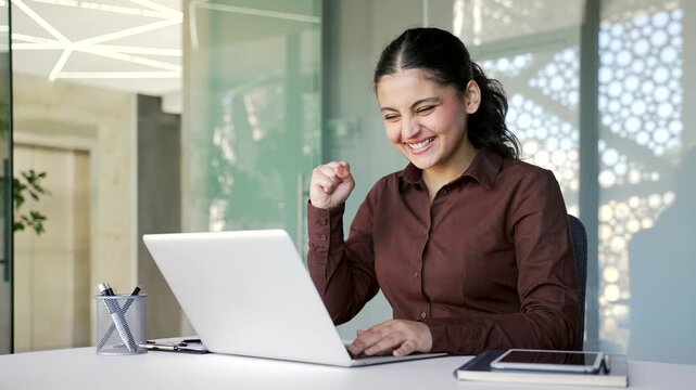 Smiling happy businesswoman reading good news on laptop sitting at workplace in business office. Satisfied glad female employee celebrates success, rejoices at positive message on computer. 