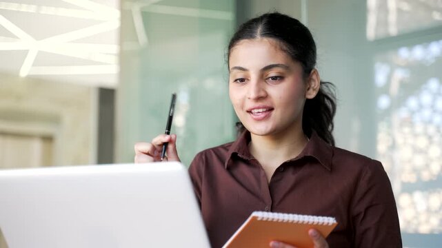 Young businesswoman watching video call online conference taking notes looking at laptop screen in office. Female worker listening remote business training, talking communicates with tutor. Close up