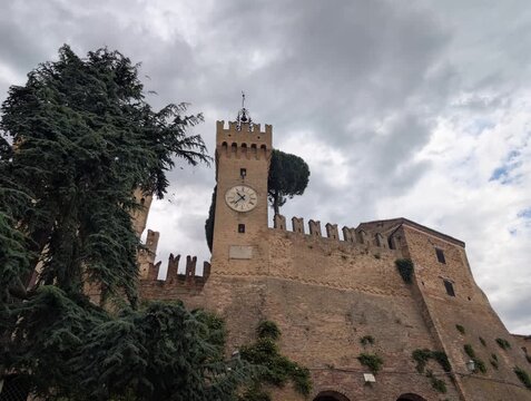 Castello storico con torre dell&rsquo;orologio, mura merlate e albero imponente sotto un cielo nuvoloso, per un&rsquo;atmosfera medievale intensa e suggestiva