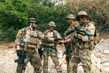 Fototapeta premium Four camouflaged soldiers in tactical gear stack hands, showing teamwork during a field operation on rocky terrain bordered by dense shrubs.