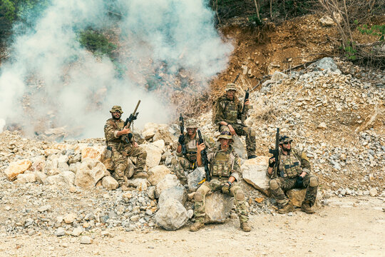 Armed soldiers in camouflage take cover among rocky terrain as smoke billows behind them during a field operation or training exercise.