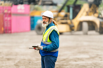 Obraz premium Construction worker in hard hat and safety vest holding a tablet at an industrial yard with containers and heavy machinery in the background.