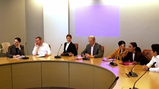 Diverse professionals seated around a curved conference table during a meeting, with microphones and documents, in a boardroom beneath a blank projector screen