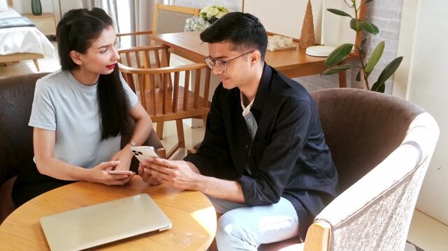 Two young adults sit at a cozy home table comparing smartphones, discussing plans, with a laptop nearby in a bright, modern living space.