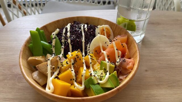 Colorful salmon poke bowl served with black rice, avocado, mango, soft-boiled egg, green vegetables and sesame seeds. Fresh healthy meal presented in a wooden bowl at a modern restaurant