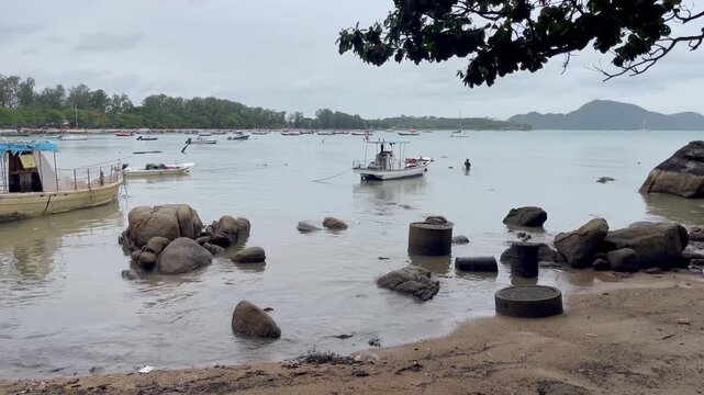 Peaceful coastal scene in Rawai, Phuket, with fishing and longtail boats anchored in shallow waters surrounded by rocky shoreline and distant hills. Authentic local maritime life in southern Thailand