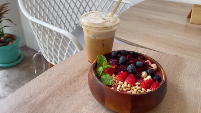 Healthy berry smoothie bowl with strawberries, blueberries, dried berries, nuts and fresh mint, served alongside iced coffee in a cozy modern caf&eacute;. Nutritious breakfast and wellness lifestyle concept
