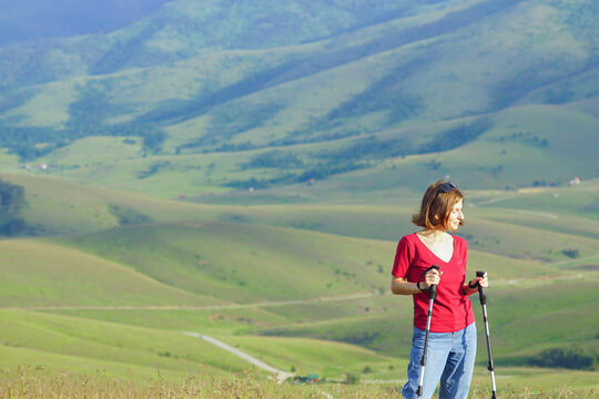 Smiling woman pauses with trekking poles on sunlit slope, broad valley spreading far below. Represents Nordic walking fitness, preventive wellness, and active outdoor recreation in mountain landscapes