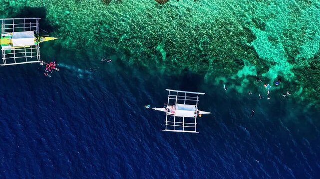 Stunning top-down drone view over Moalboal, Philippines. Features traditional outrigger boats anchored on a dramatic coral reef drop-off filled with snorkelers and divers in clear turquoise water.
