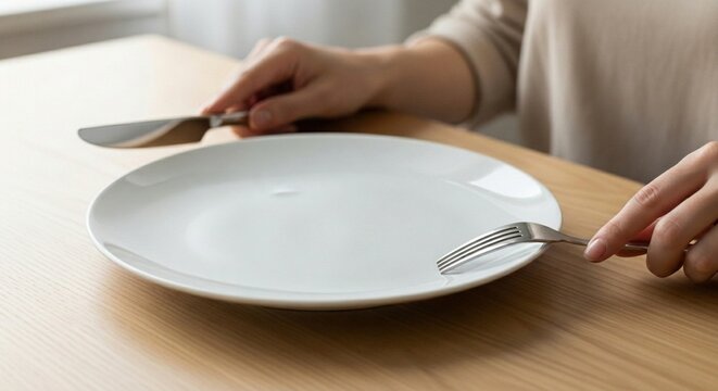 Empty white plate with knife and fork held by person at wooden dining table representing hunger, dieting or waiting for food.