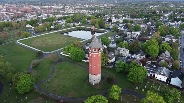 Historic red brick water tower rising from green park with reflecting pond, residential blocks and urban skyline with church spires stretching to horizon. Massachusetts, USA. Aerial orbit shot.