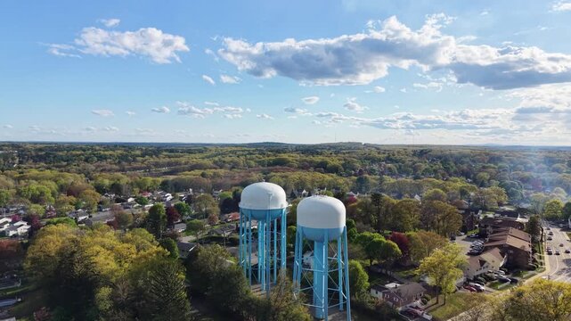 Two blue water towers rising above dense autumn tree canopy, suburban neighborhood and busy road visible through fall foliage under blue sky with white clouds. Massachusetts, USA. Aerial approachingx