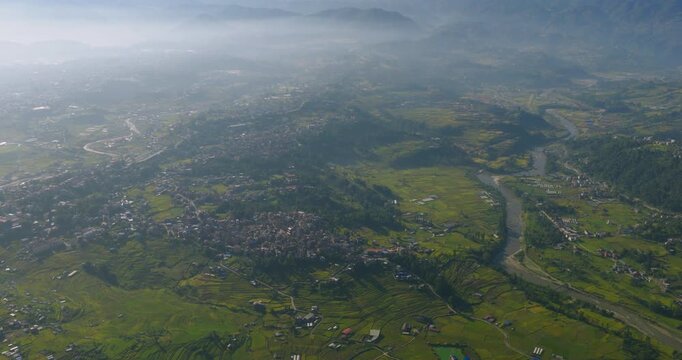 Drone moves in a top-down view over a wide valley, showing green terrace farming fields, a winding river, a rural settlement, and misty mountains in Pharping, Nepal