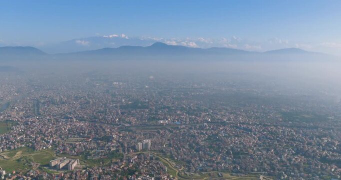 Drone moves sideways in a lateral tracking view over the dense city landscape of the Kathmandu Valley, Nepal with the snow-covered Himalayan mountain range visible in the distant background
