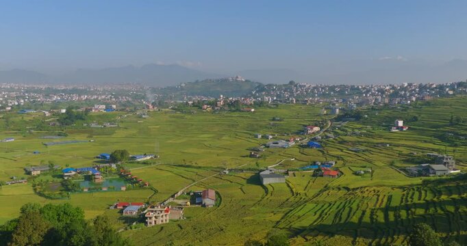 Drone glides forward over a wide landscape of green agricultural terrace fields, scattered village buildings, and hills under a clear blue sky in Pharping, Nepal, agricultural land and greenery