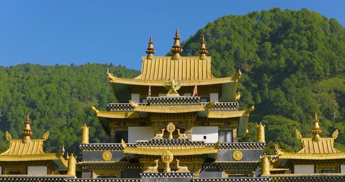 Drone pull out from a multi-tiered Tibetan Buddhist monastery building characterized by its ornate golden tiered roofs, situated against a green hillside backdrop in Pharping, Nepal