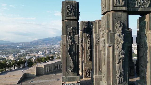 Drone aerial view of the famous Chronicles of Georgia monument columns in Tbilisi, showcasing monumental bronze sculptures with a panoramic city landscape background. High quality 4k footage