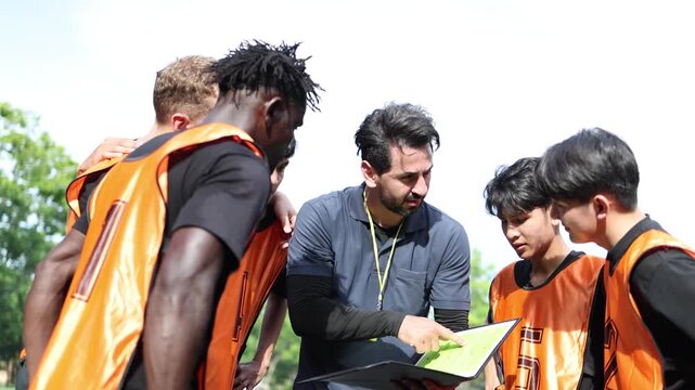 A football coach points to a playbook while discussing strategy with a diverse group of male soccer players during a team huddle on an outdoor field in daylight