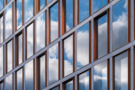 Clouds reflect across a blue glass facade, where windows, architecture, building grid and business surfaces create mirrored urban depth