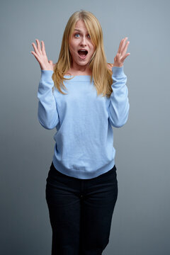 Cheerful woman with a surprised expression, wearing a blue sweater and black pants, against a plain background. Perfect for concepts of joy, celebration, astoundment, and positive emotional reactions.