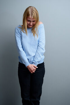A young woman wearing a light blue sweater and black pants smiles shyly while looking downwards, conveying a sense of modesty and introspection. The sene has a positive and reflective mood.