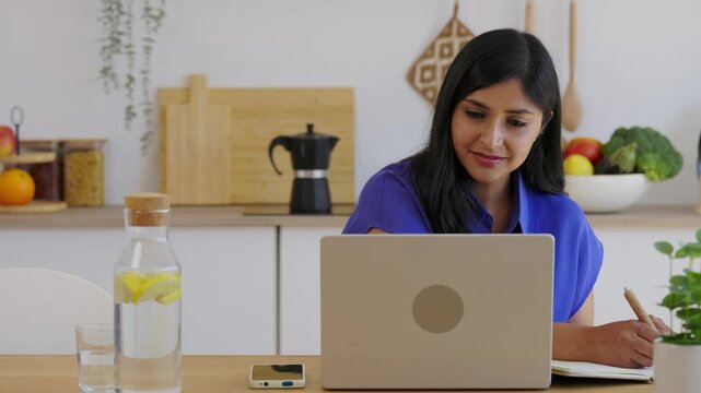 Mid adult Indian woman taking notes during online course. Female learner writing study notes while following digital training on laptop. Education and professional development concept