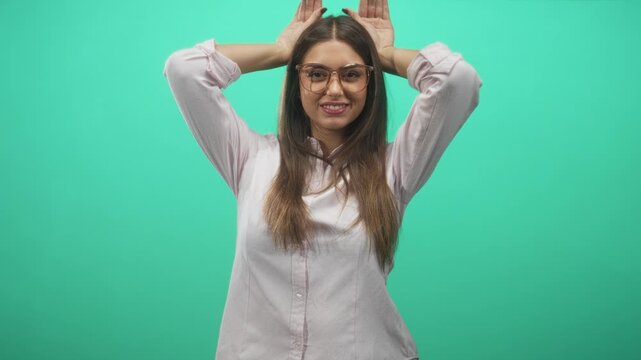Young woman in glasses raises hands behind head making rabbit ears gesture against a mint green backdrop in studio; playful energy.