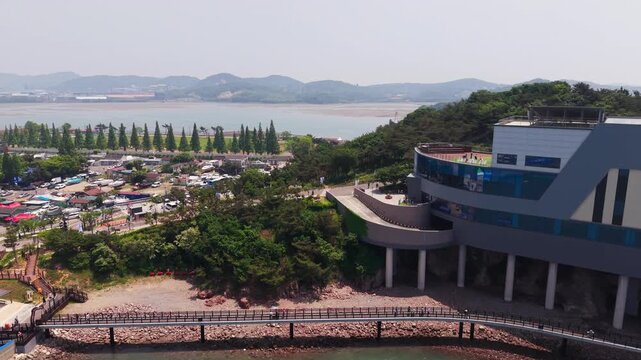 Aerial forward toward Jebudo Marine Cable Car Seohaerang Jeongok Station modern building with tourists walking on seaside promenade above coastal park in South Korea
