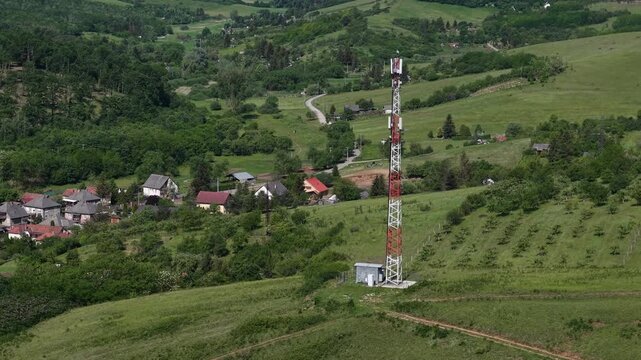 A tall cell tower stands on a grassy hillside overlooking a small rural village in a lush green valley.