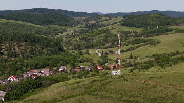 A cell tower stands tall in a vast green landscape, overlooking a small rural village under a clear summer sky.