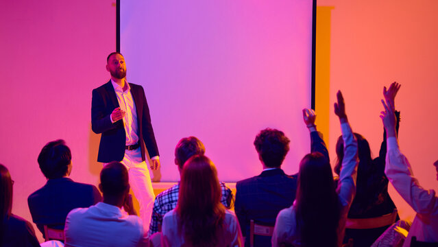 Male speaker answering audience questions beside blank screen in neon seminar room. Concept of Q and A session, business workshop, leadership training, public speaking and corporate education.