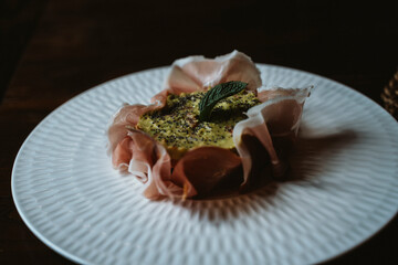 Tartlet topped with creamy avocado, sprinkled with quinoa, surrounded by delicate prosciutto, and garnished with a mint leaf, presented on a white textured plate. © Nadezda Anokhina