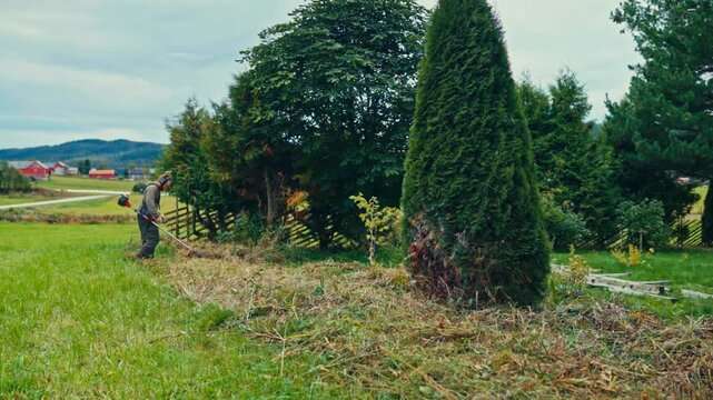 A Man is Clearing Overgrown Grass and Bushes Near a Garden Fence in a Rural Landscape - Timelapse