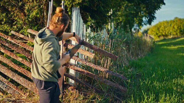 A Man is Sawing a Wooden Fence Outdoors Along a Grassy Field - Timelapse