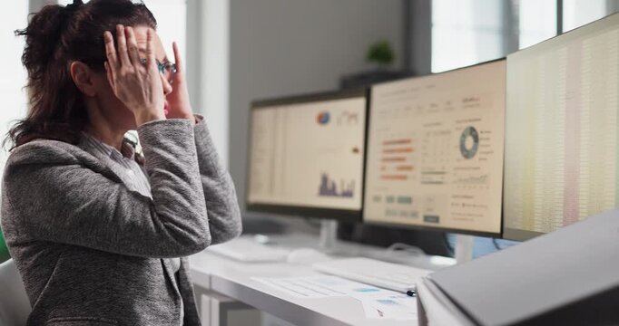 Stressed woman analyst in office rubbing headache. Dashboards on computer screens show complex reports and tight deadlines, driving fatigue, pressure, and overwork. Concept of workplace stress.
